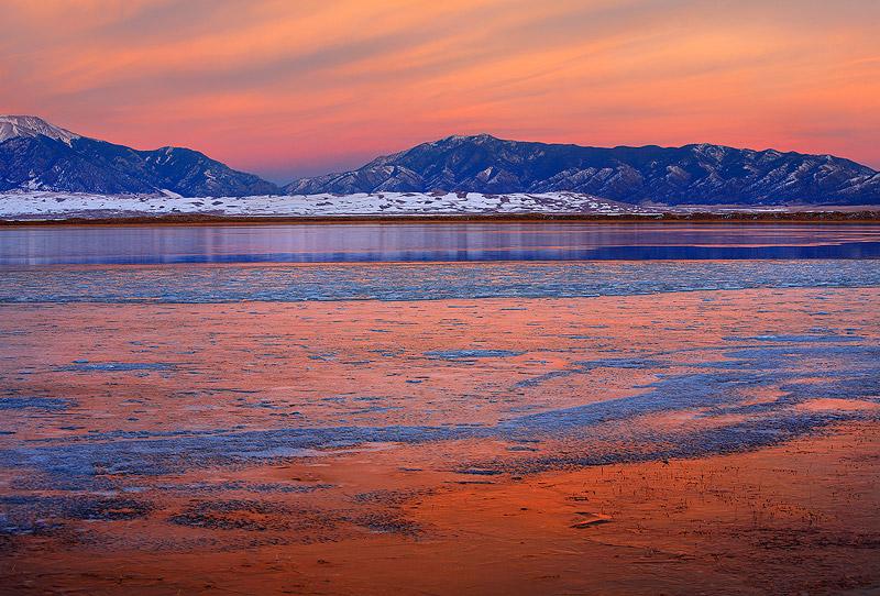 Frozen lake reflects a fiery sunset. dunes in the distance.